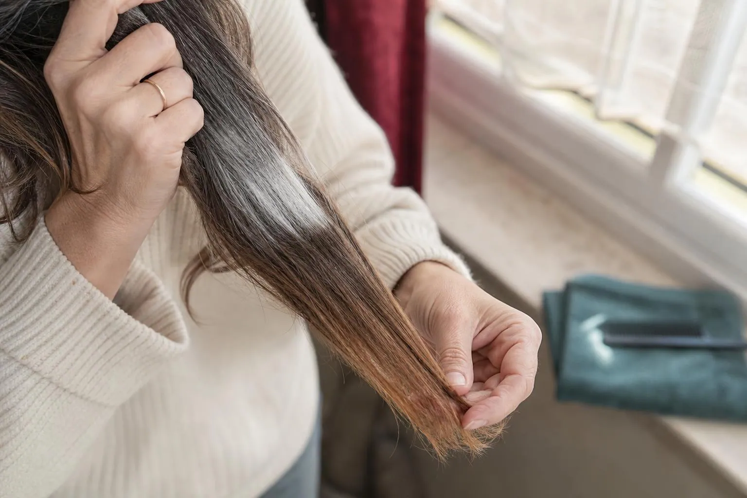 Gros plan sur des cheveux mi longs tenus à deux mains devant une fenêtre, montrant une racine blanche, des longueurs châtain et des pointes plus claires